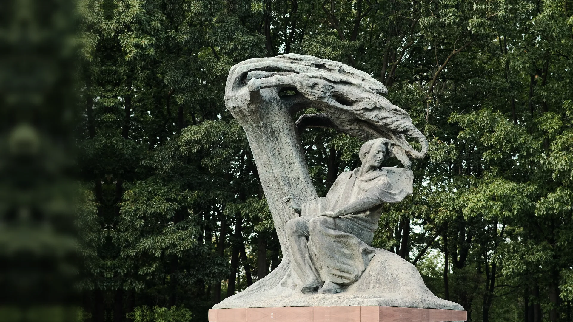The bronze Chopin Monument in Warsaw's Łazienki Park: the composer seated beneath a sweeping willow-shaped canopy of metal, with green trees behind.